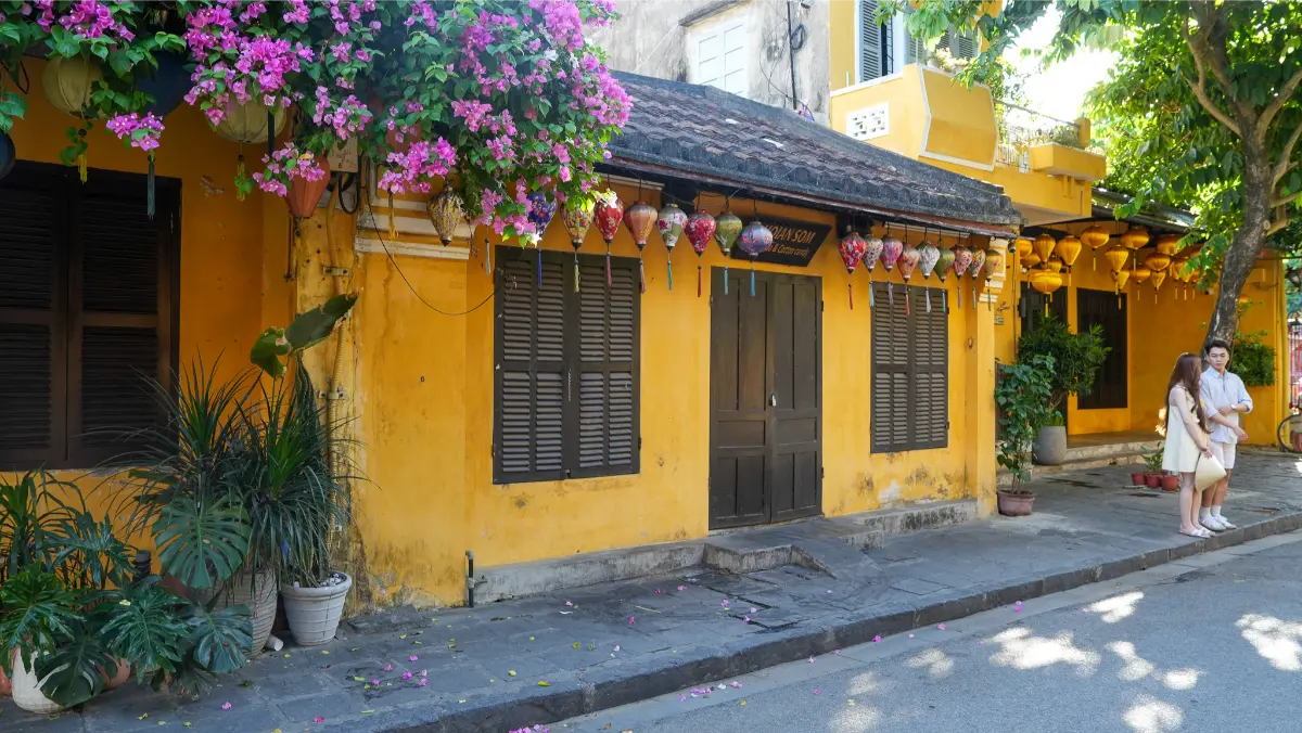 Pink bougainvillea cascading over a yellow Hoi An heritage house with lanterns and a young couple standing by the entrance.