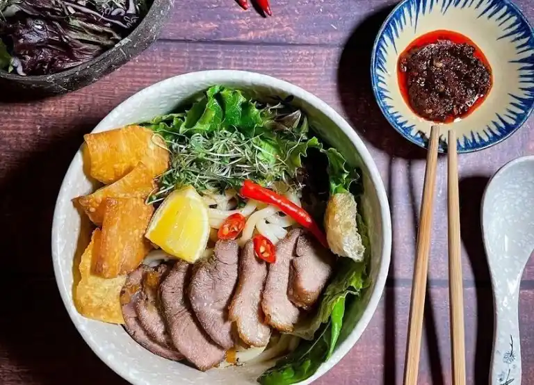Bowl of traditional cao lầu noodles in Hoi An with pork, greens, crispy rice crackers and herbs.