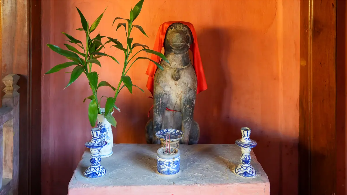 Stone guardian monkey statue draped in a red cloth on a small altar inside the Japanese Covered Bridge in Hoi An, with bamboo stems and blue-and-white porcelain candlesticks.