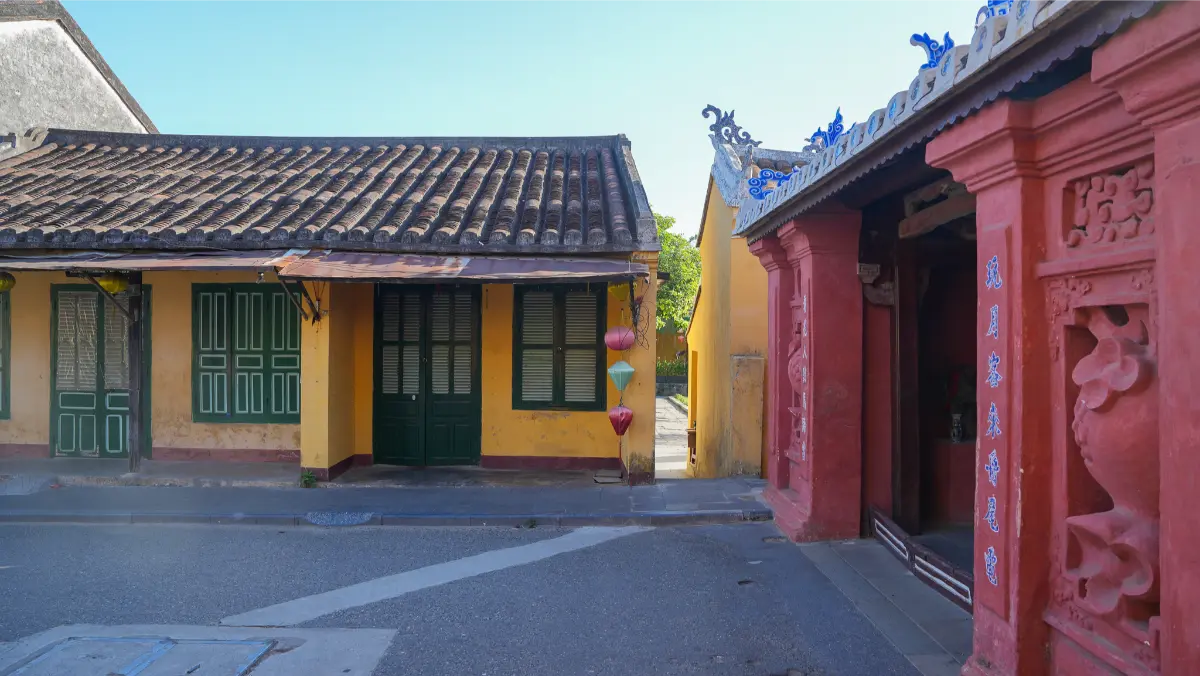 Traditional yellow heritage house with green shutters and terracotta-tiled roof standing next to the red walls of the Japanese Covered Bridge in Hoi An Ancient Town.