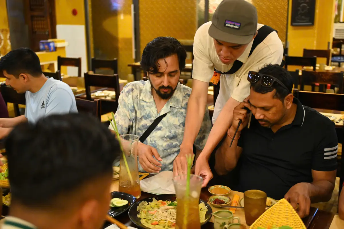 Local Hoi An guide explaining a bowl of cao lau noodles to travellers seated at a warm-lit restaurant table.