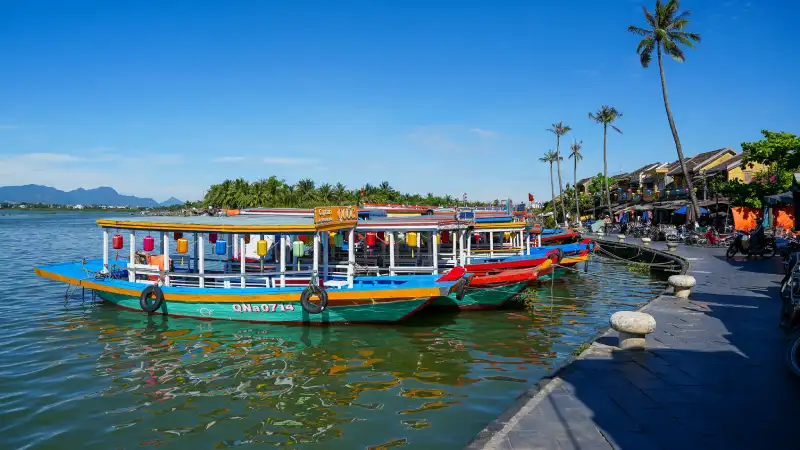 Traditional wooden boats drifting along the Thu Bồn River in Hoi An Ancient Town at golden hour, framed by ochre-yellow heritage houses.