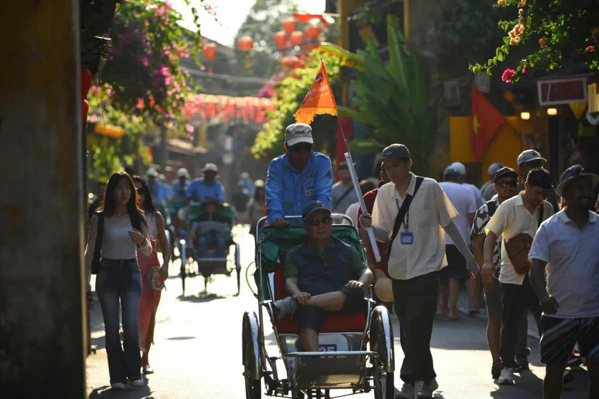 Traveller riding a traditional cyclo through a lantern-lined street of Hoi An Old Town while the local guide walks alongside with an orange flag.