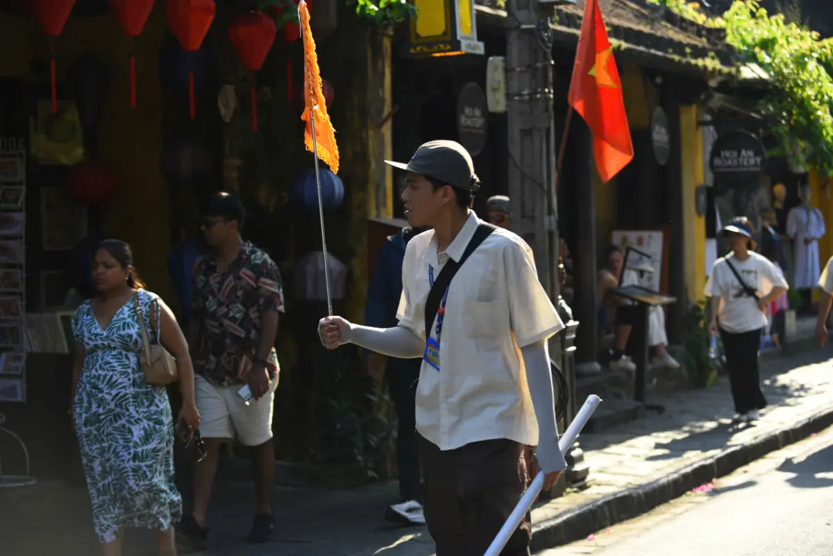 Young local guide holding an orange Free Walking Tours Hoi An flag on a sunlit street lined with red lanterns and a Vietnamese flag.