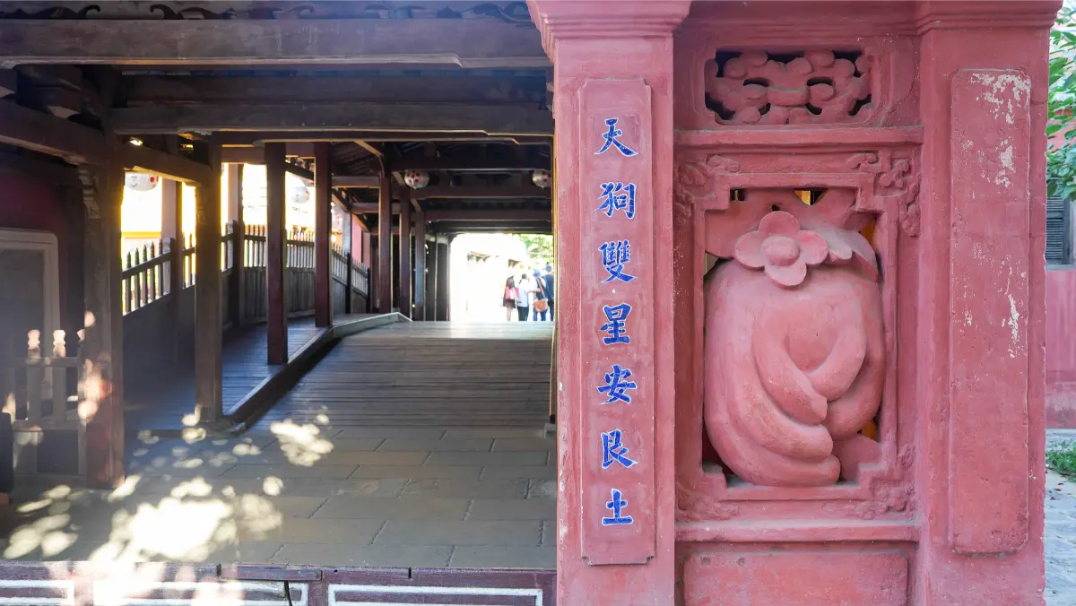 View from inside the Japanese Covered Bridge in Hoi An looking through the red-pillared doorway, with a carved peach relief and Chinese inscriptions framing the wooden walkway.