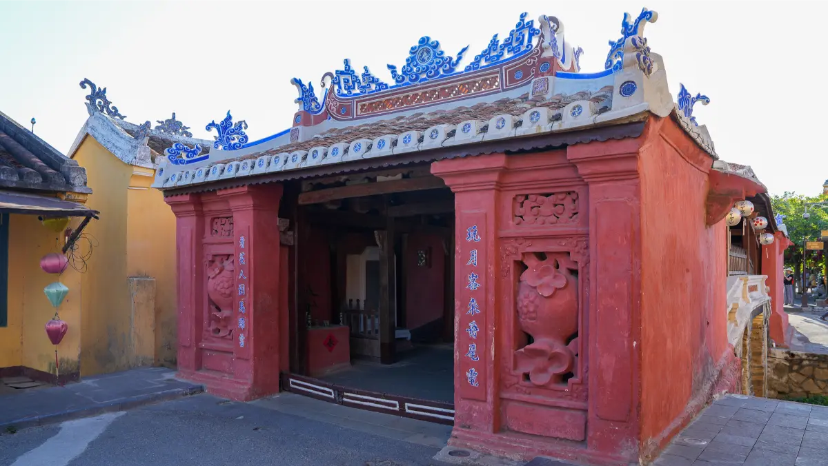 East entrance of the Japanese Covered Bridge in Hoi An, showing carved red pillars, ornate blue and white roof cresting and pastel silk lanterns hanging beside the doorway.