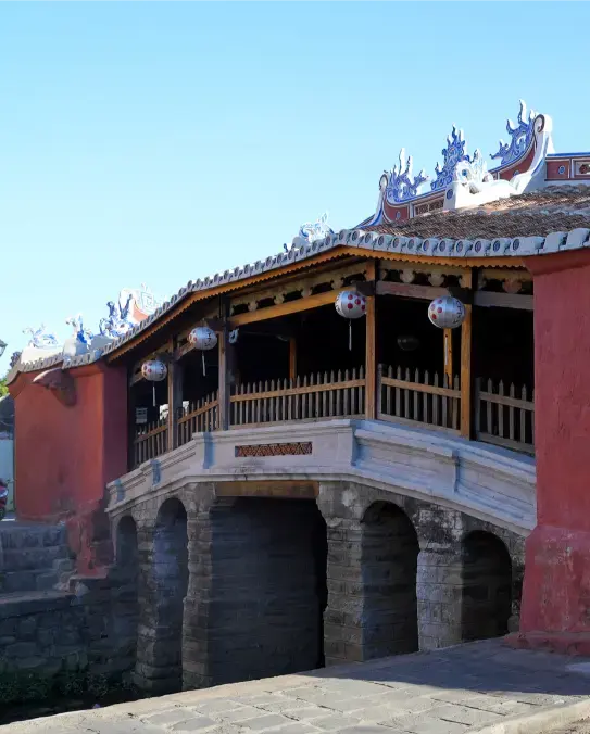 Japanese Covered Bridge in Hoi An Ancient Town seen from the southwest, showing its pink walls, tiled roof and elegant wooden structure under a clear blue sky.