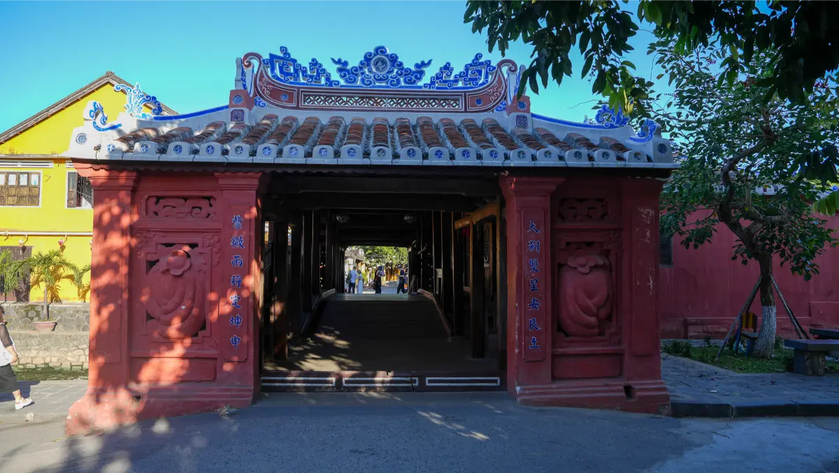 Main entrance of the Japanese Covered Bridge in Hoi An framed by yellow heritage walls, with intricate blue and white roof carvings above the red-pillared gateway.