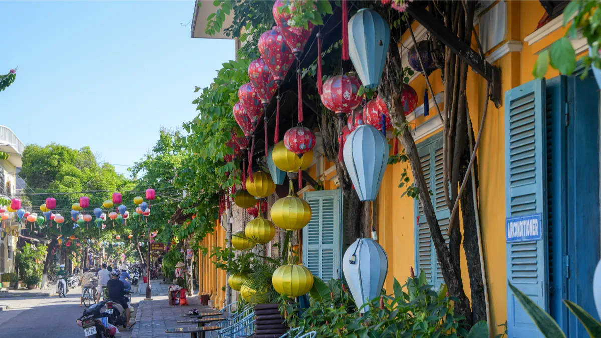 Colourful silk lanterns hanging in front of a yellow Hoi An shophouse with blue shutters, stretching down a lantern-lined street.