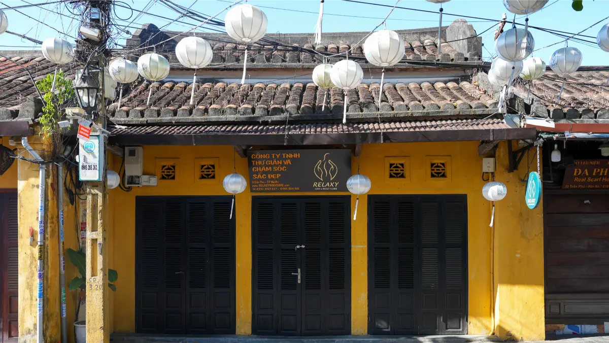 Traditional yellow Hoi An house with a tiled roof, white lanterns hanging above dark wooden shutters and Vietnamese signage.