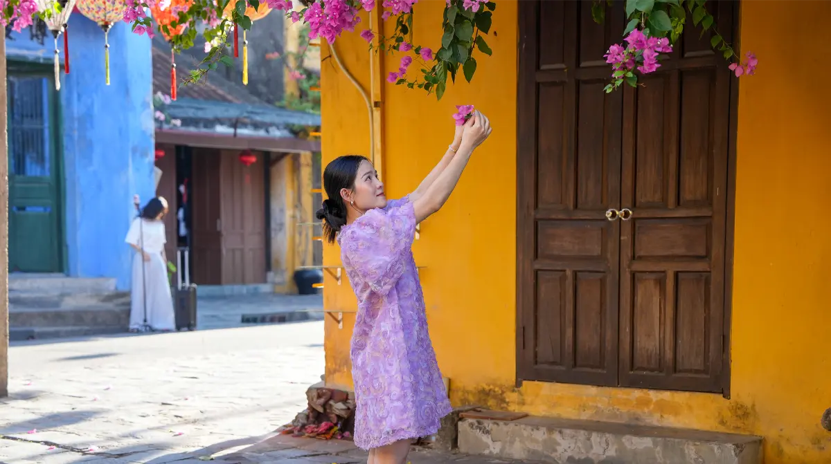 A young woman in a lilac áo dài reaches up to bougainvillea blooms beside a bright yellow heritage wall in Hoi An Ancient Town.