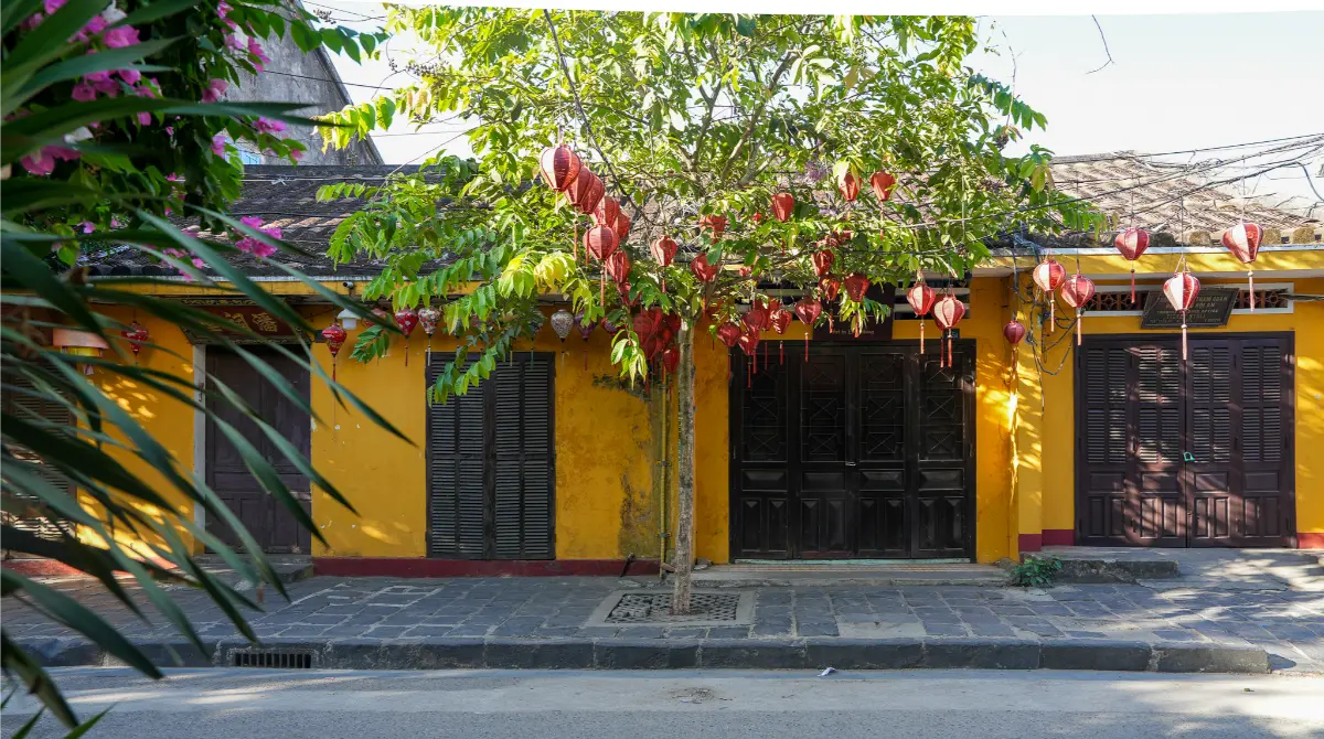 Ochre-yellow heritage shophouse in Hoi An Ancient Town with red silk lanterns hanging from a leafy tree and dark wooden shutters.
